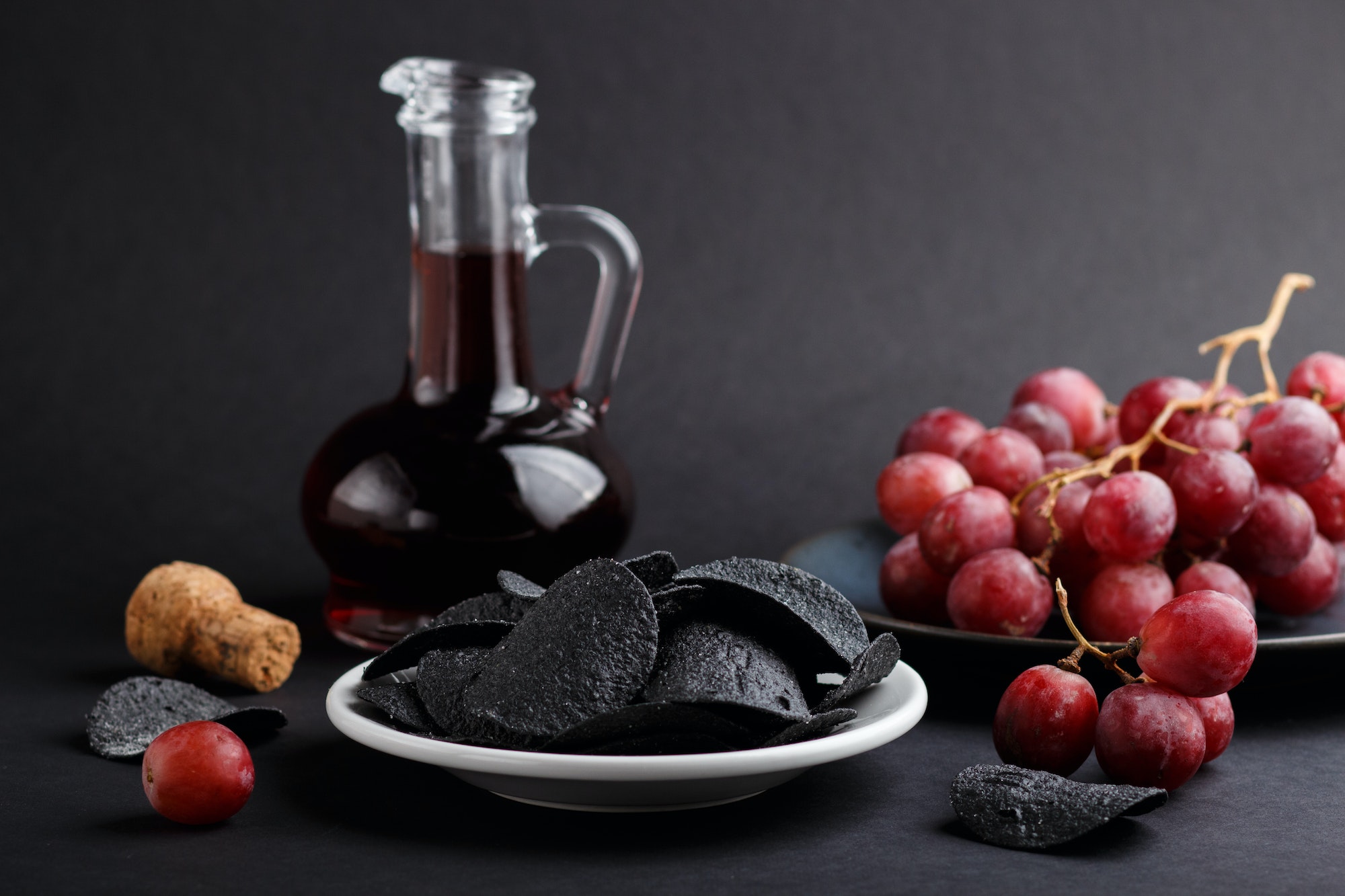 Black potato chips with charcoal, balsamic vinegar in glass, red grapes on a blue ceramic plate